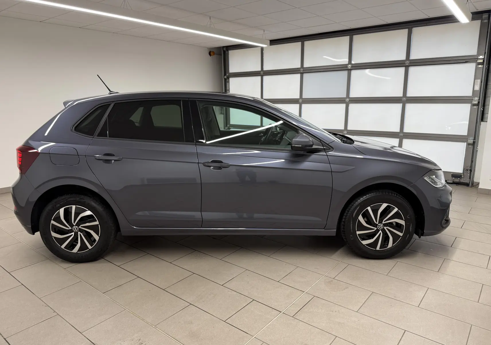 Profil droit d'une Volkswagen Polo gris cendre 2025 avec jantes alliage bicolores et vitres surteintées en intérieur lumineux.
