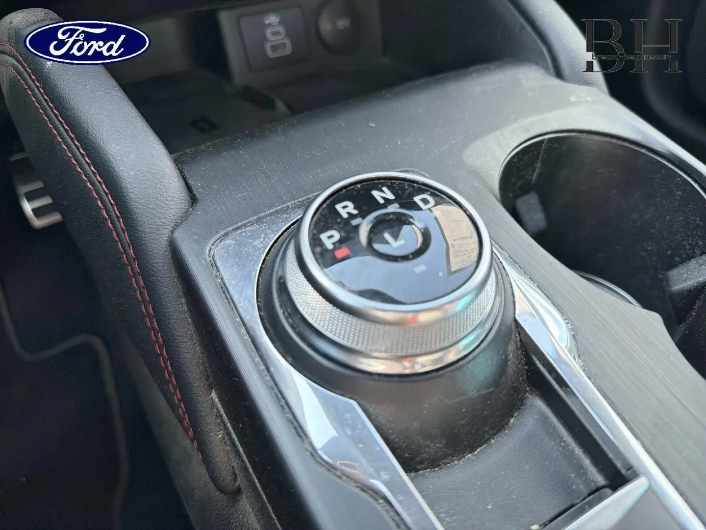 Gros plan sur la molette de sélection de vitesses et l'accoudoir central en cuir noir avec surpiqûres rouges dans un Ford Kuga 2024.