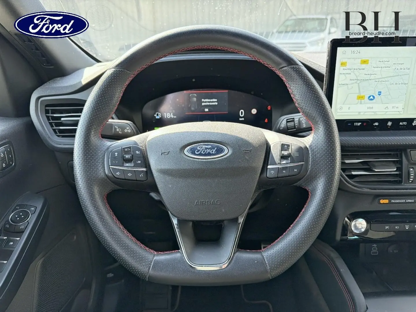 Vue intérieure centrée sur le volant sport noir à surpiqûres rouges du Ford Kuga 2024 avec tableau de bord numérique et écran tactile GPS.