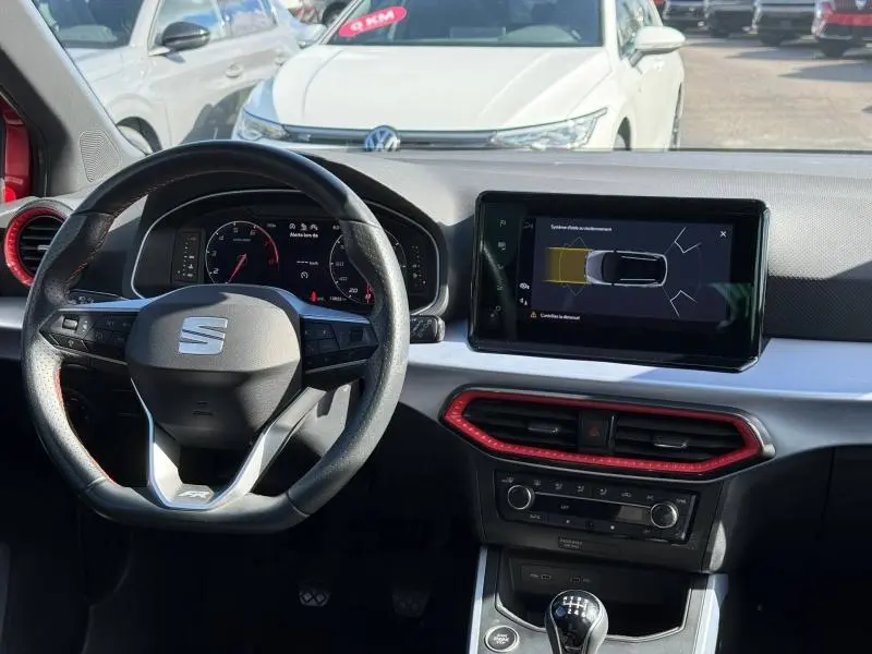 Vue intérieure du poste de conduite du SEAT Arona 2025 avec volant multifonction, écran tactile et détails rouges sur la console.