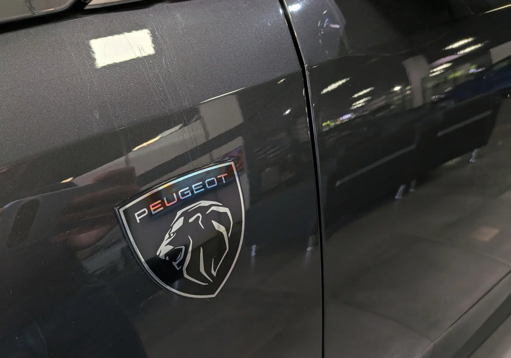 Gros plan sur l’écusson Peugeot noir et gris sur la porte gris titane d’un 5008 hybride 2025.