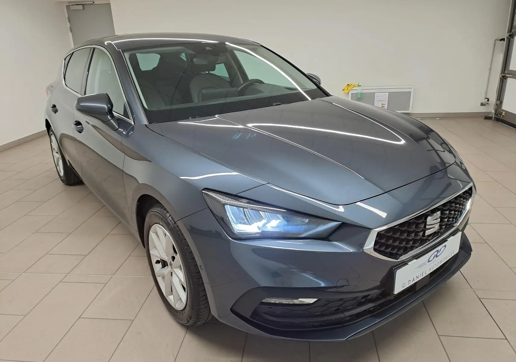 SEAT Leon 1.5 eTSI Hybrid gris magnétique vue 3/4 avant en intérieur, phares allumés et vitres surteintées arrière.