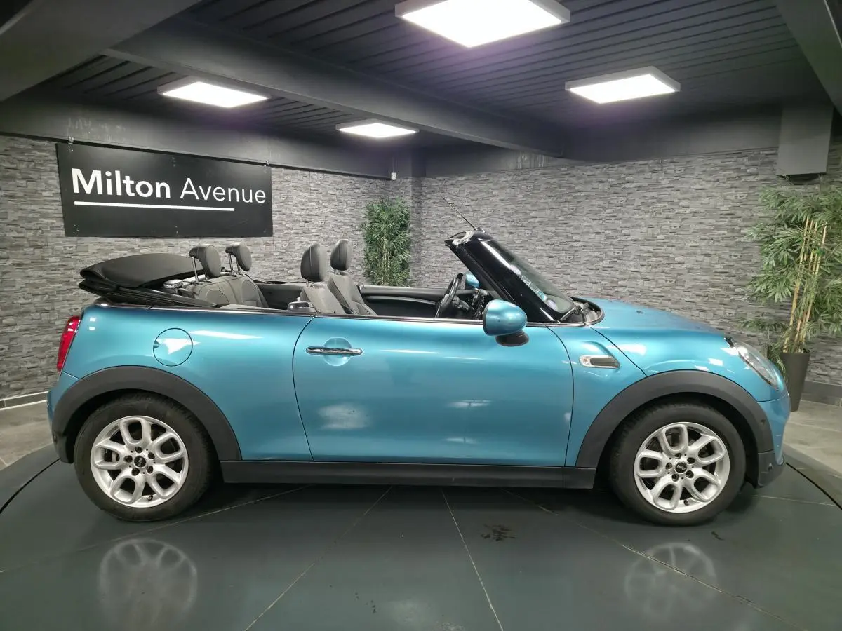 Vue latérale droite d'une MINI Cabriolet bleu avec capote noire repliée et jantes argentées en intérieur showroom.