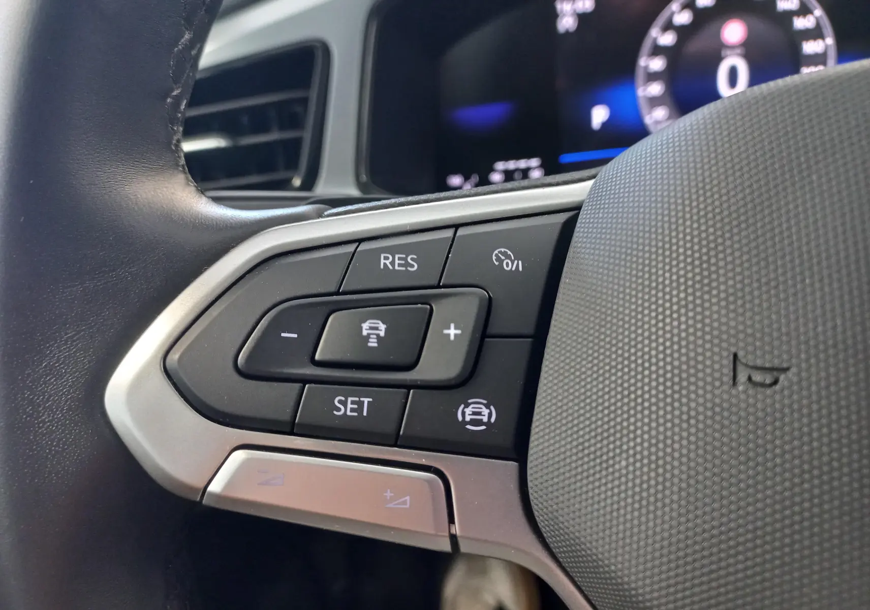 Gros plan sur les commandes au volant côté gauche du Volkswagen T-Roc 2025 gris Indium, avec affichage numérique en arrière-plan.