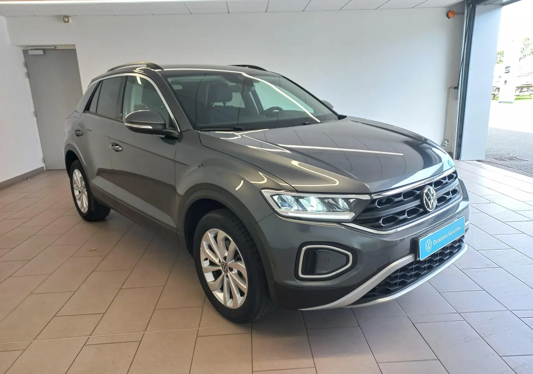 Volkswagen T-Roc 2025 gris Indium en vue 3/4 avant droit, avec calandre distinctive et jantes alliage visibles.