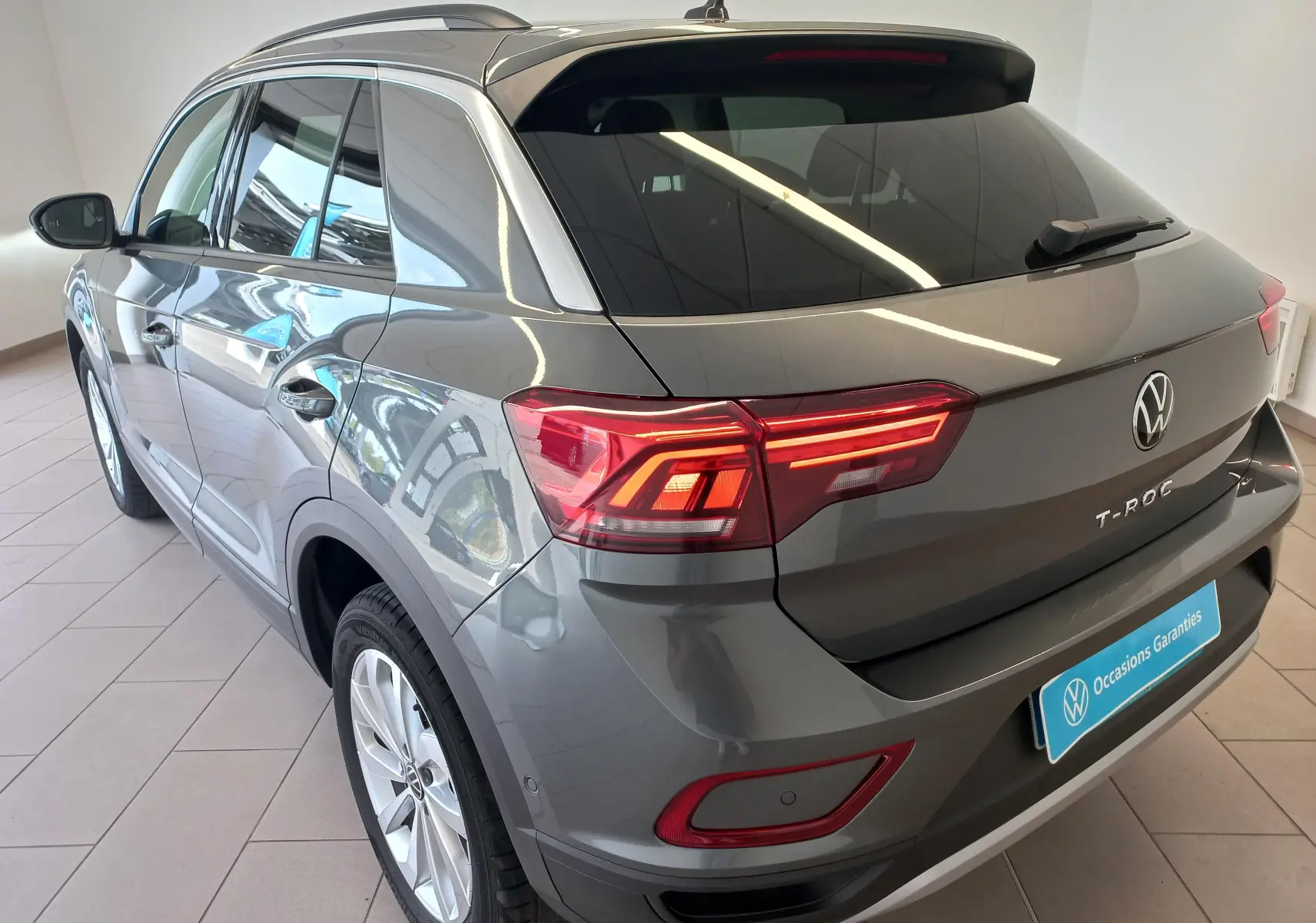 Vue 3/4 arrière droite d'un Volkswagen T-Roc gris Indium 2025 avec feux arrière LED et toit noir brillant.