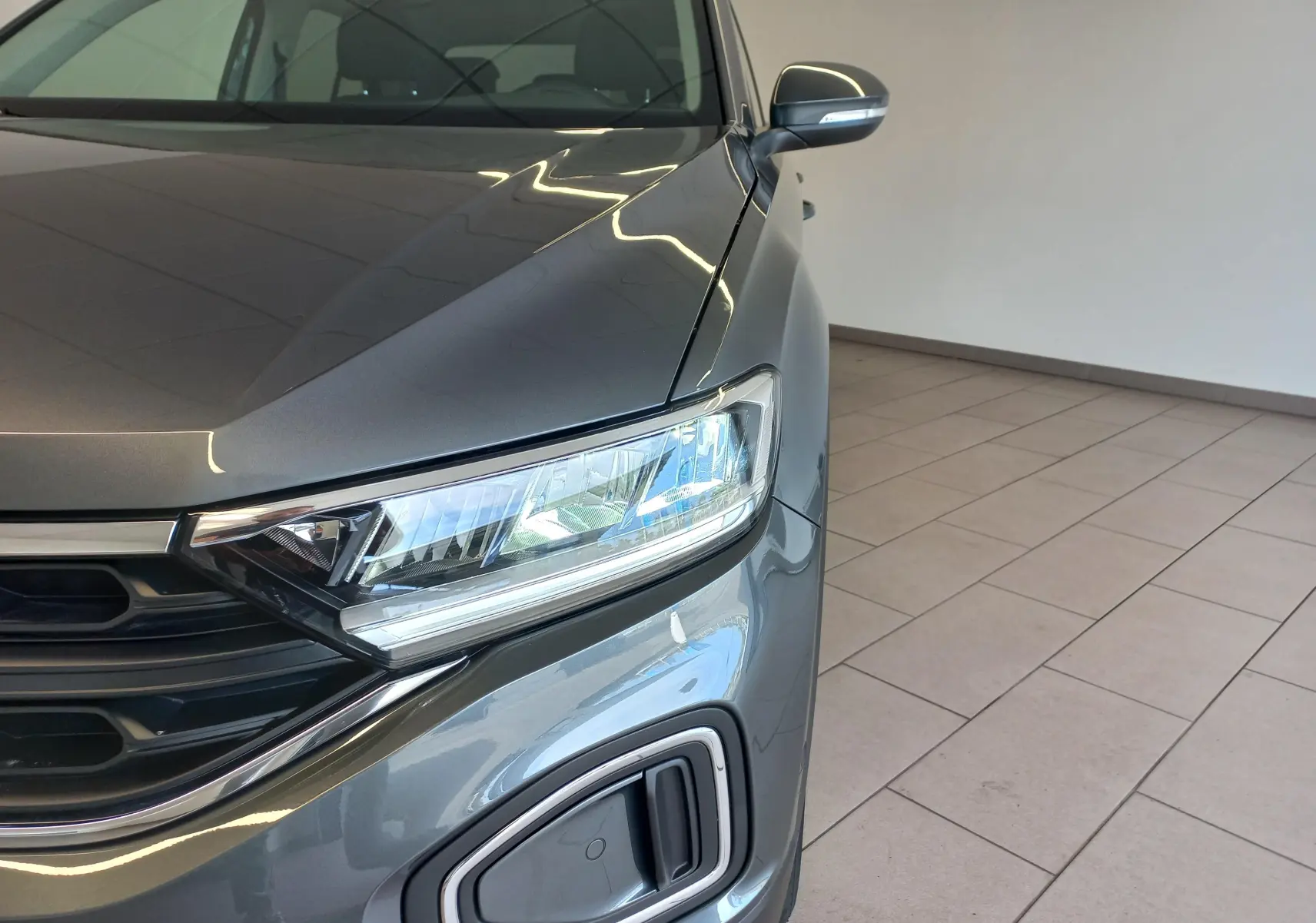 Gros plan sur l'avant droit du Volkswagen T-Roc 2025 gris Indium, mettant en valeur le phare et la calandre moderne.
