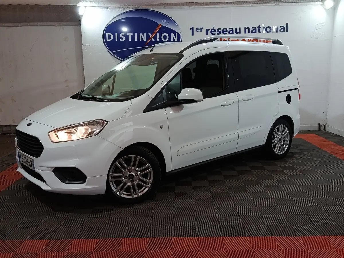 Vue 3/4 avant droite d’un Ford Grand Tourneo Connect blanc avec phares allumés dans un showroom intérieur.