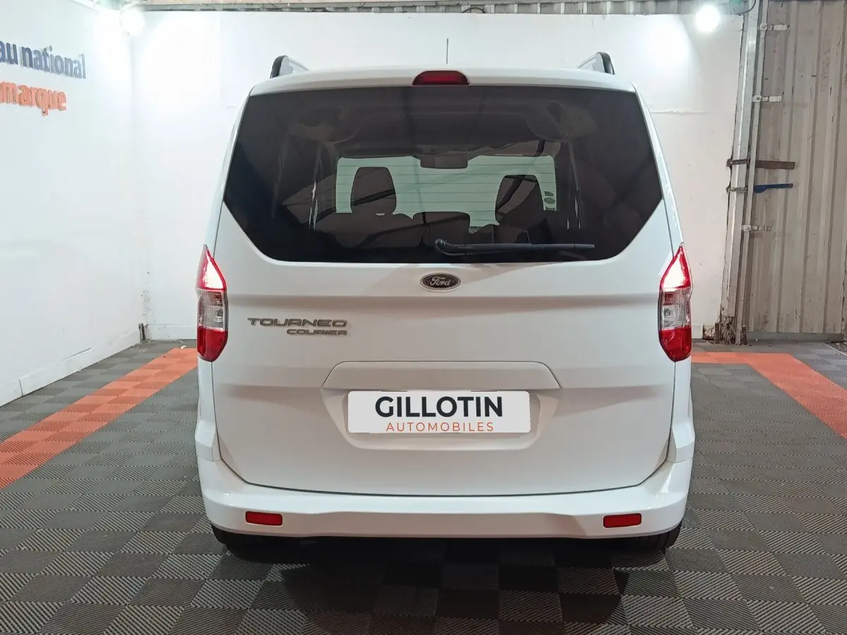 Vue arrière d’un Ford Grand Tourneo Connect blanc, avec feux arrière allumés et logo visible sur un sol à damier.