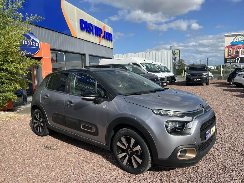 Citroën C3 gris vue 3/4 avant droit, avec toit noir et protections latérales caractéristiques sur parking extérieur.