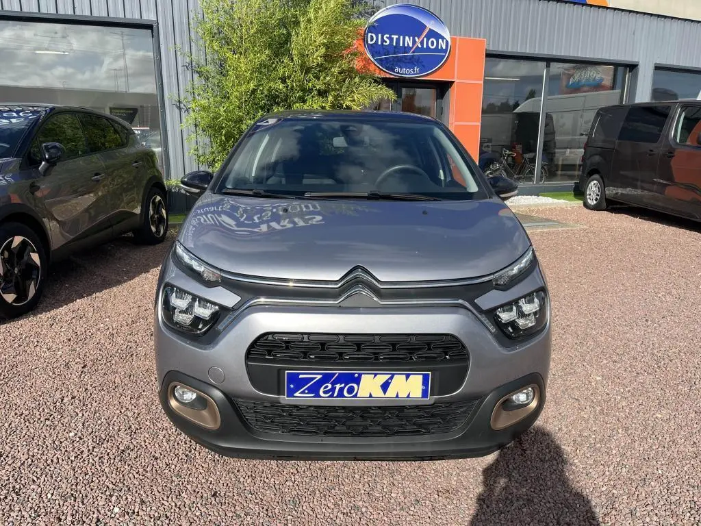 Vue frontale d'une Citroën C3 gris métallisé avec phares LED et plaque Zéro KM sur parking extérieur.