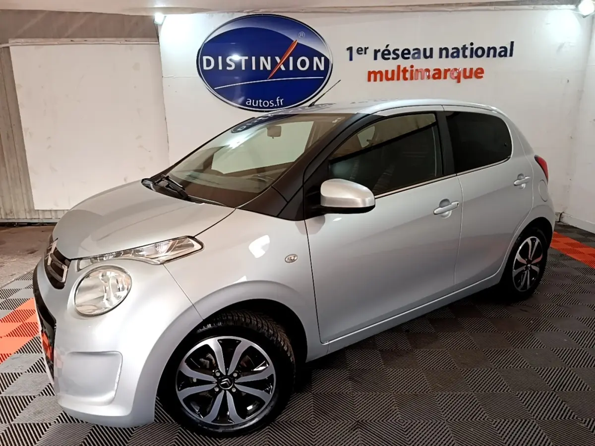Citroën C1 gris clair vue 3/4 avant droit en intérieur, avec jantes noires et logo Distinxion en arrière-plan.