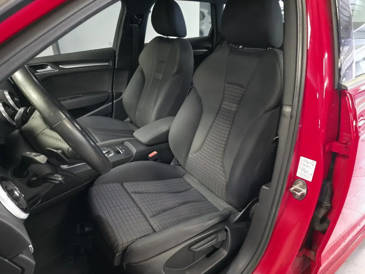 Vue intérieure côté conducteur de l'Audi A3 Sportback rouge, montrant les sièges avant en tissu et cuir noir.