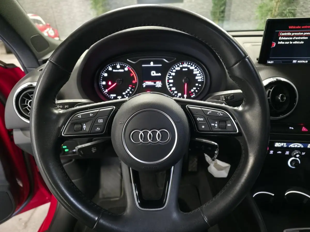 Vue intérieure centrée sur le volant noir multifonction de l'Audi A3 Sportback rouge avec tableau de bord et écran central visibles.