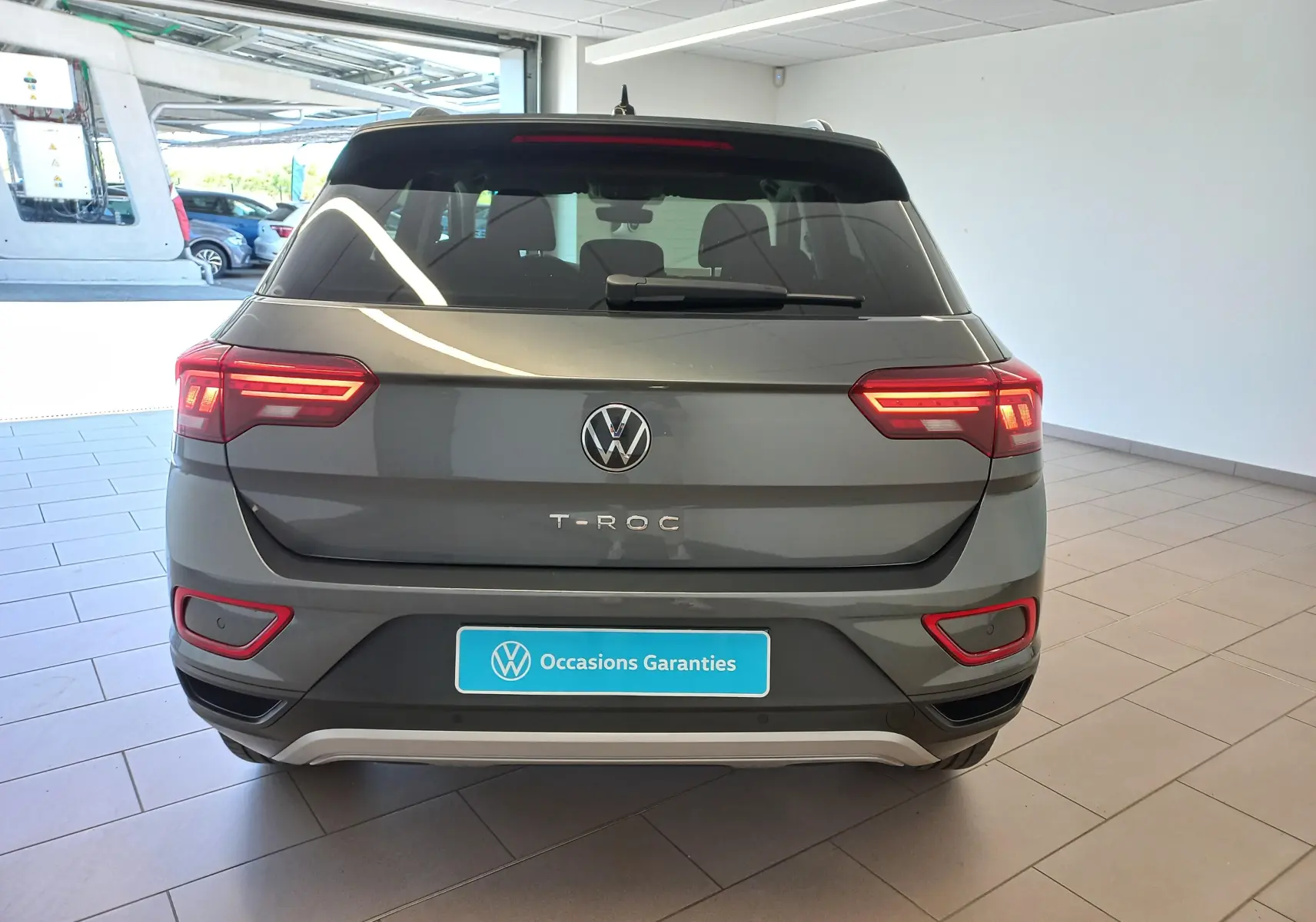 Vue arrière d'un Volkswagen T-Roc 2025 gris Indium avec feux allumés et plaque "Occasions Garantis" en intérieur.