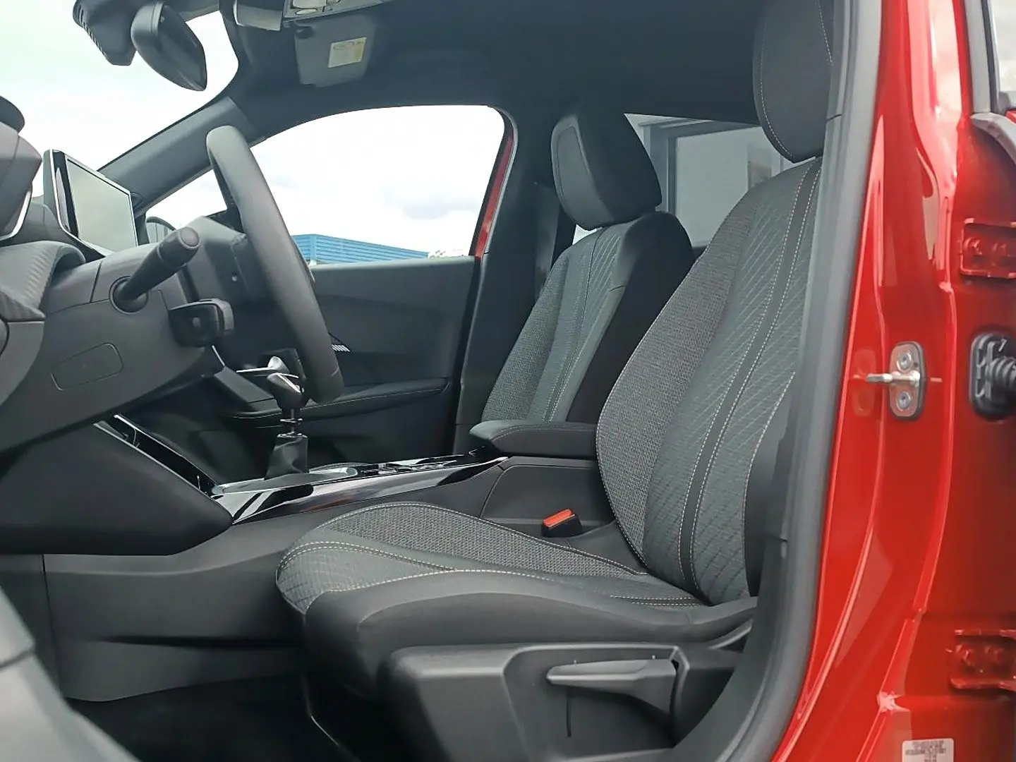 Vue intérieure côté conducteur du Peugeot 2008 rouge elixir, montrant les sièges en tissu gris et la console centrale.