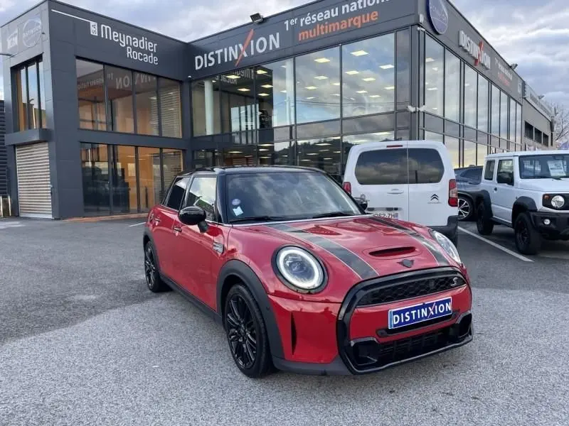 Vue 3/4 avant droite d'une MINI 5 portes rouge avec bandes noires sur le capot, roues noires, devant un showroom Distinxion.