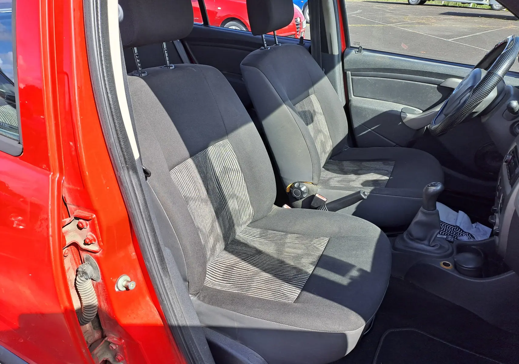 Vue intérieure côté conducteur de la Dacia Sandero rouge 2010, sièges tissu noir avec motifs rayés et levier de vitesse manuel.