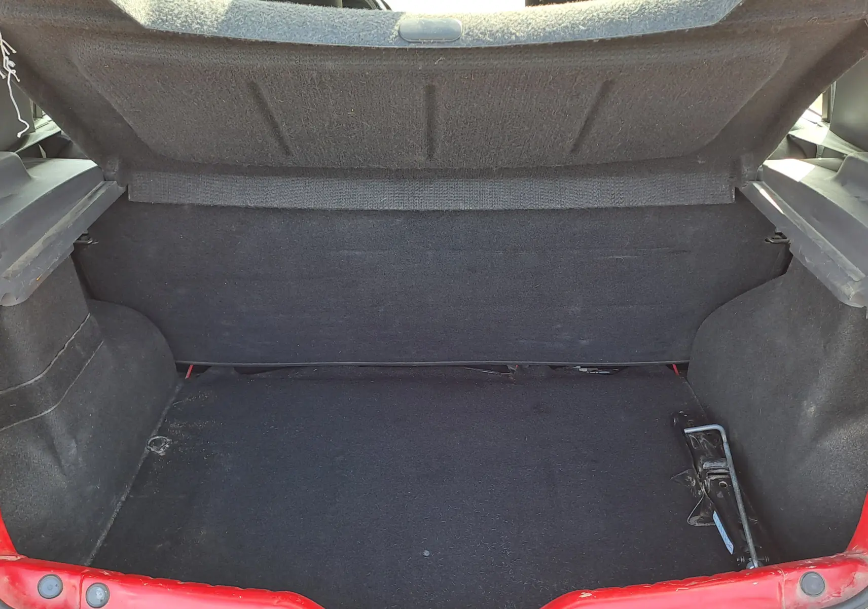 Coffre ouvert d'une Dacia Sandero rouge 2010, vue arrière montrant l'espace de chargement vide et le cric côté droit.