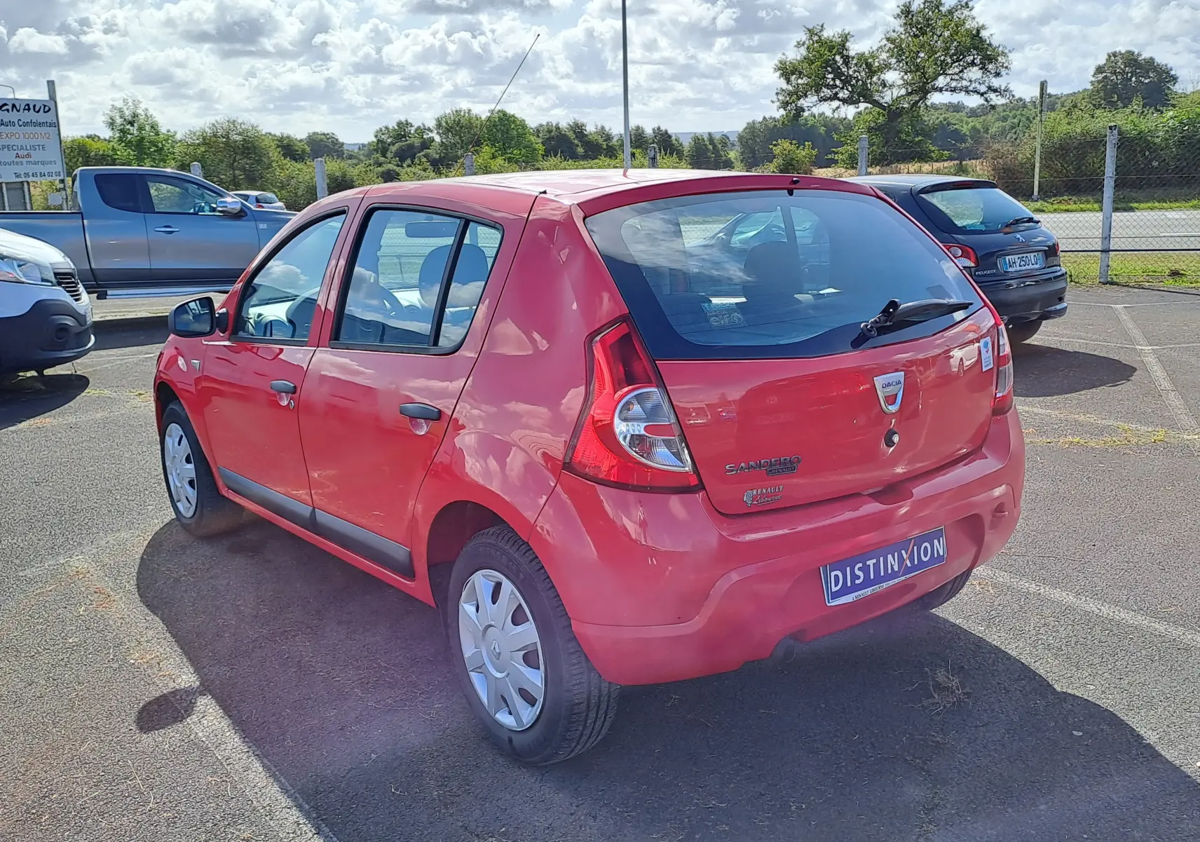 Vue 3/4 arrière droite d'une Dacia Sandero rouge de 2010 avec plaques et badges visibles sur un parking extérieur.