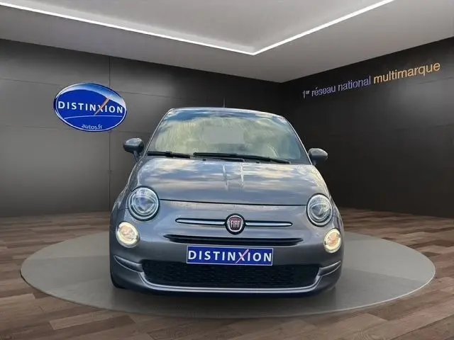 Vue avant d'une FIAT 500 gris foncé avec phares ronds et logo rouge, sur un podium intérieur showroom.
