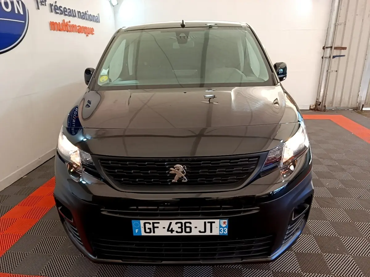 Vue avant d’un Peugeot Partner noir 2022 avec calandre noire et phares allumés dans un garage.