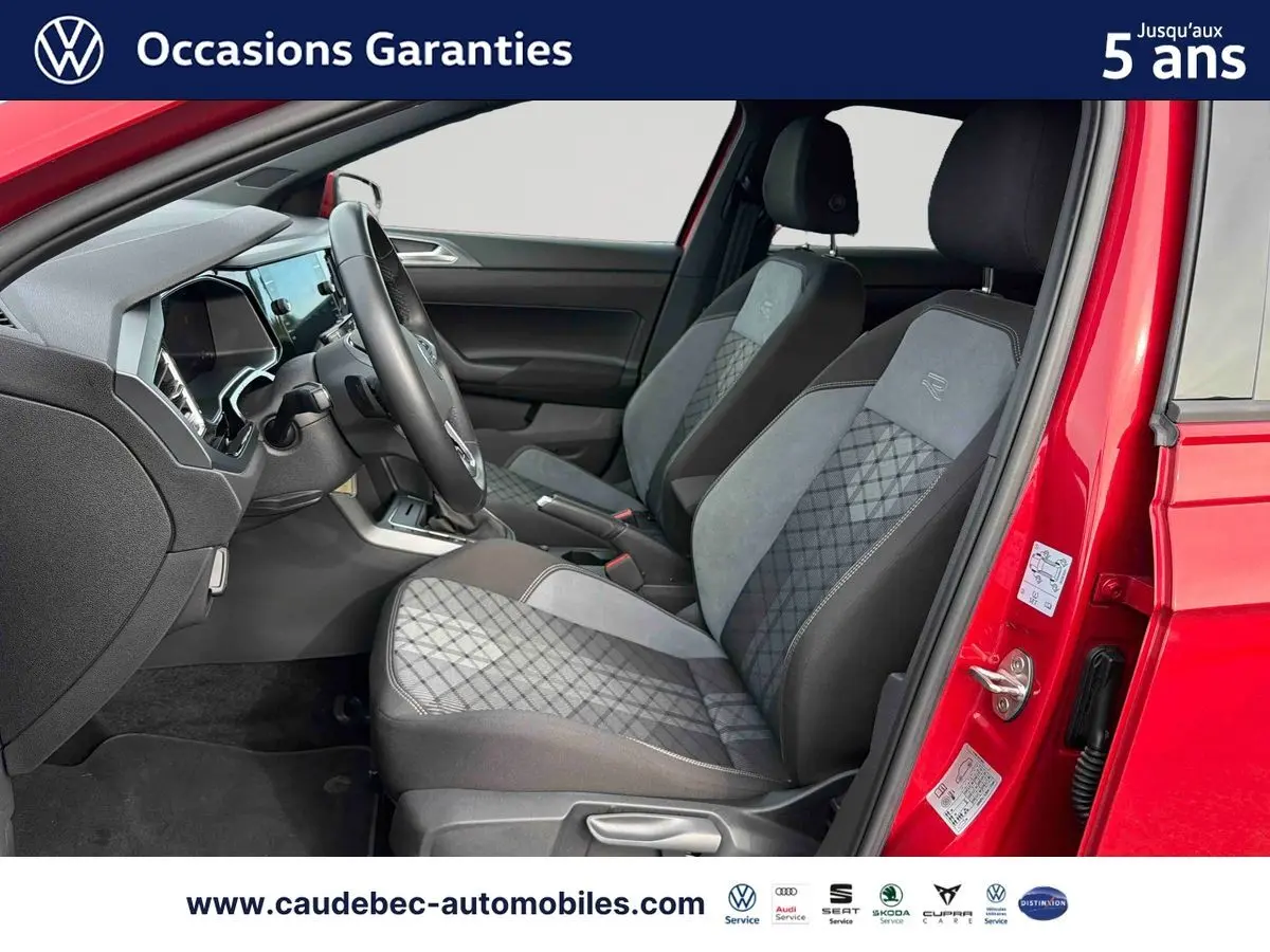 Vue intérieure côté conducteur d'une Volkswagen Polo rouge 2025, sièges avant tissu gris à motifs et volant cuir multifonctions.