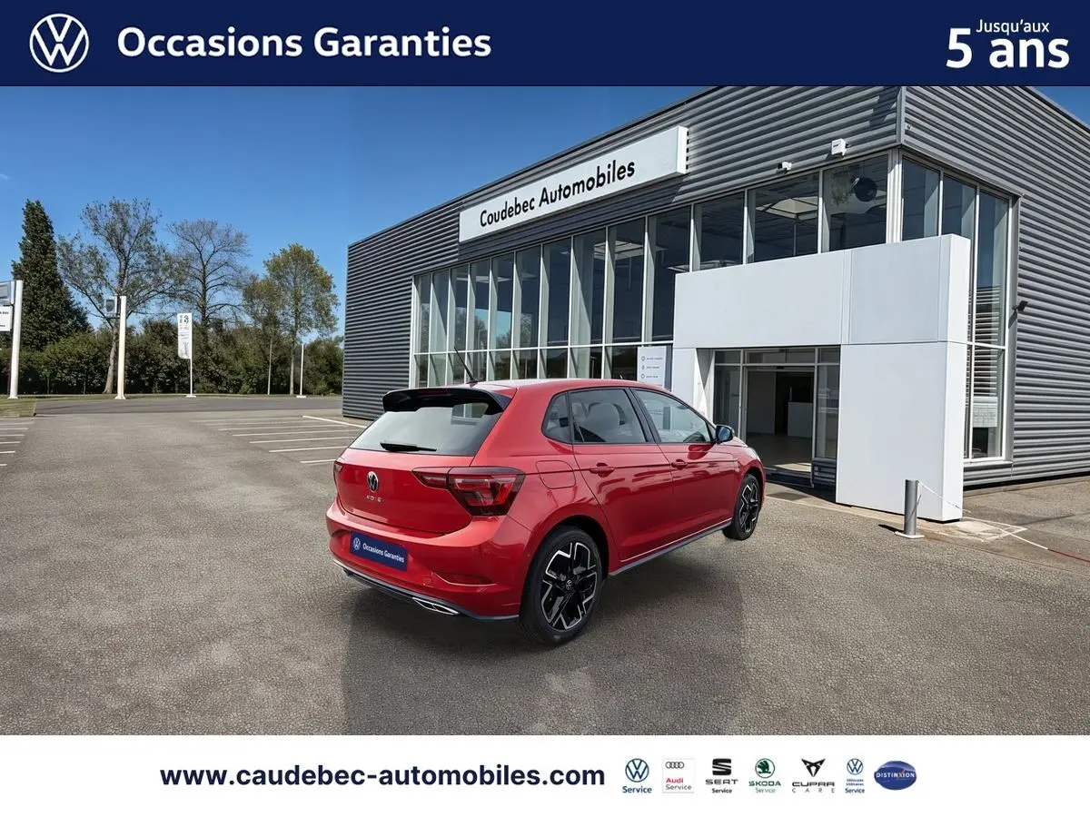 Vue 3/4 arrière droite d'une Volkswagen Polo rouge garée devant un bâtiment moderne de concession automobile.