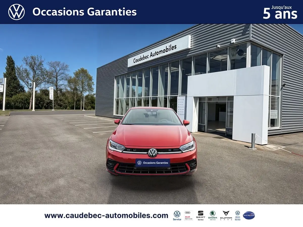 Vue avant d'une Volkswagen Polo rouge garée devant un bâtiment moderne de concession automobile Caudebec Automobiles.