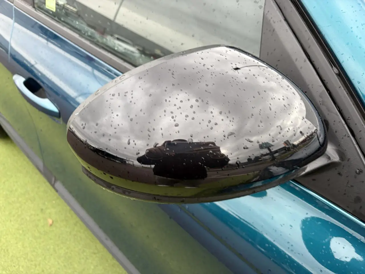 Gros plan sur le rétroviseur noir brillant côté gauche d’un Renault Arkana bleu Zanzibar métallisé avec gouttes de pluie.