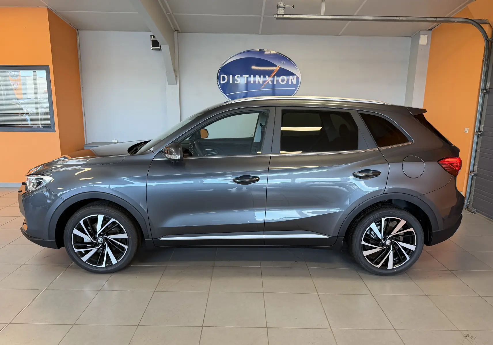 Vue de profil côté gauche du MG ZS 1.5 L Hybrid+ gris Candem avec jantes alliage 18 pouces en intérieur showroom.