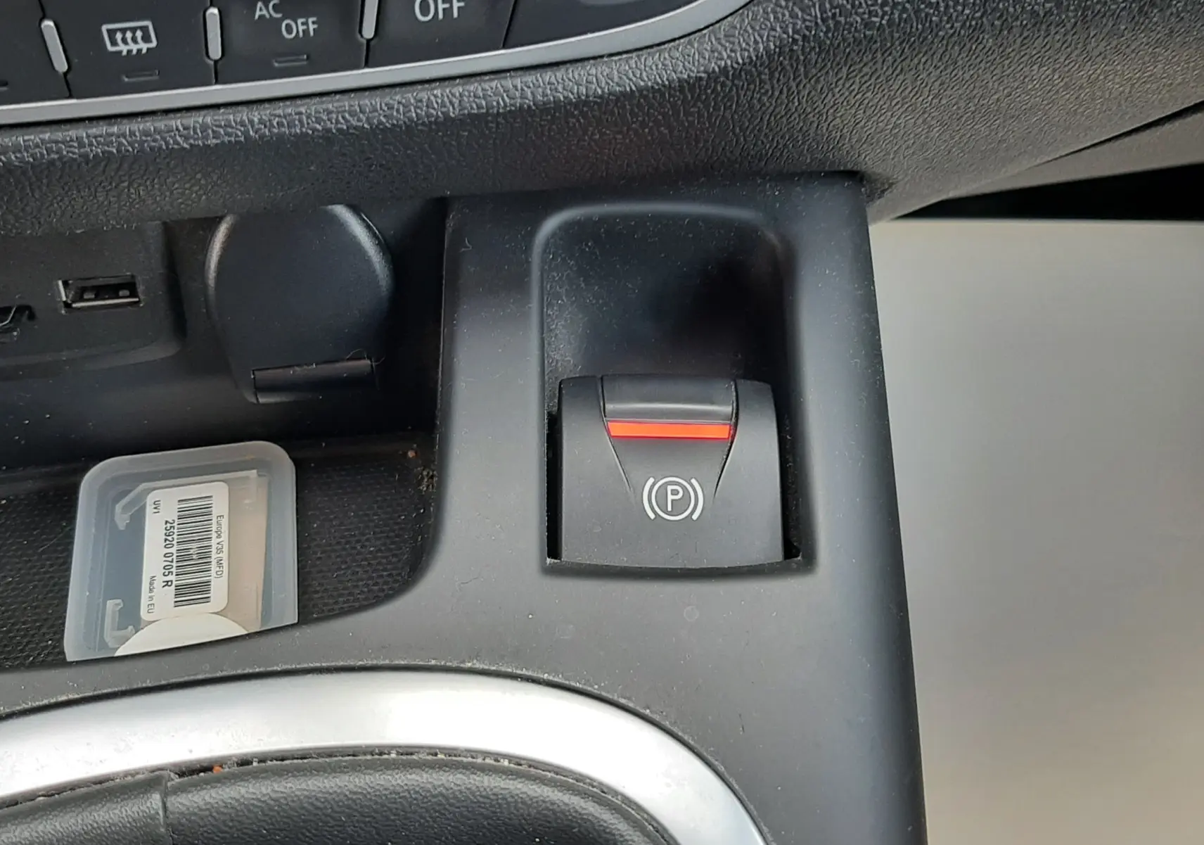 Gros plan sur le bouton de frein à main électrique noir avec témoin rouge, tableau de bord et console centrale du Renault Grand Scenic noir.