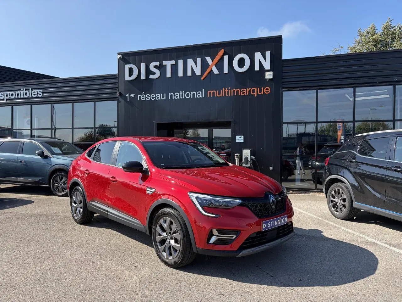 Renault Arkana rouge flamme métal vu en 3/4 avant droit, avec jantes alliage grises 17 pouces, stationné devant un showroom.