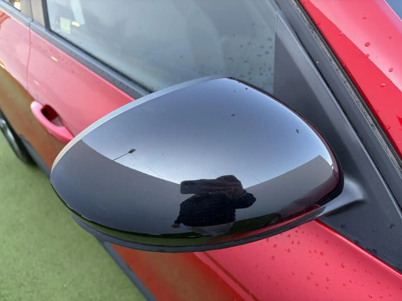 Rétroviseur noir brillant côté gauche d’un Renault Arkana Rouge Flamme Metal avec portière et poignée visibles.