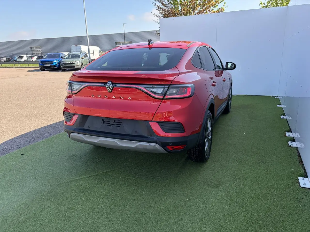 Vue 3/4 arrière droite d'une Renault Arkana rouge flamme métal avec feux arrière LED et jantes alliage grises sur sol vert.