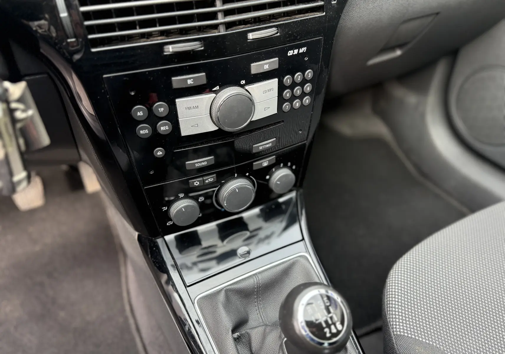 Gros plan sur la console centrale noire et le levier de vitesses manuel de l'Opel Astra gris, avec commandes radio et climatisation visibles.
