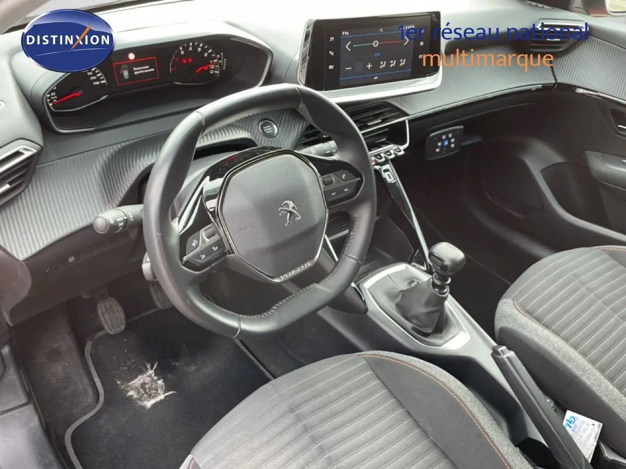 Vue intérieure centrée sur le volant et la console centrale d'une Peugeot 208 rouge Elixir, boîte manuelle visible.