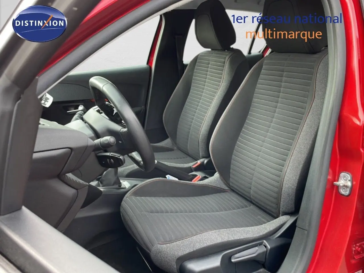 Vue intérieure côté conducteur de la Peugeot 208 rouge Elixir Metal, montrant les sièges tissu noir et gris et le volant.