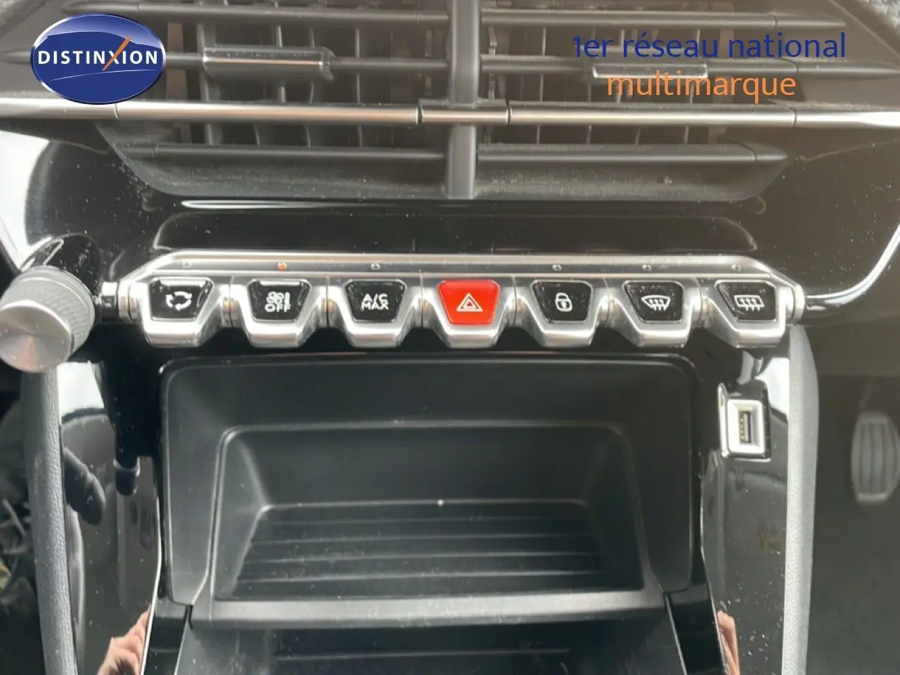 Gros plan sur la console centrale noire brillante de la Peugeot 208 rouge, avec rangée de boutons chromés et port USB visible.