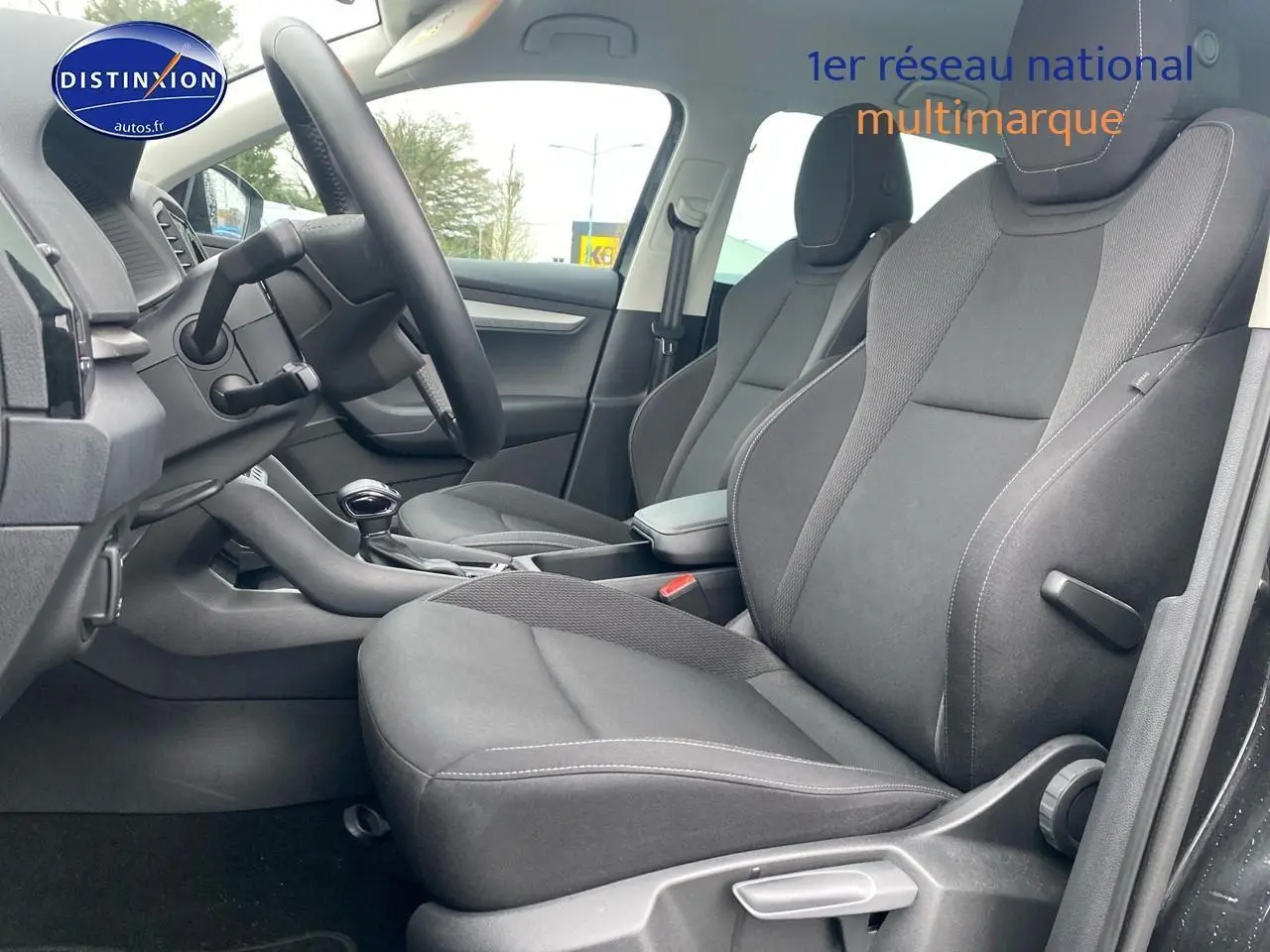 Vue intérieure côté conducteur du Skoda Karoq noir 2025, sièges tissu gris avec accoudoir central et volant cuir.