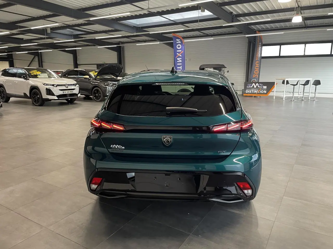 Vue arrière d'une Peugeot 308 GT bleu métallisé avec feux arrière 3 griffes LED et bandeau noir brillant en showroom.