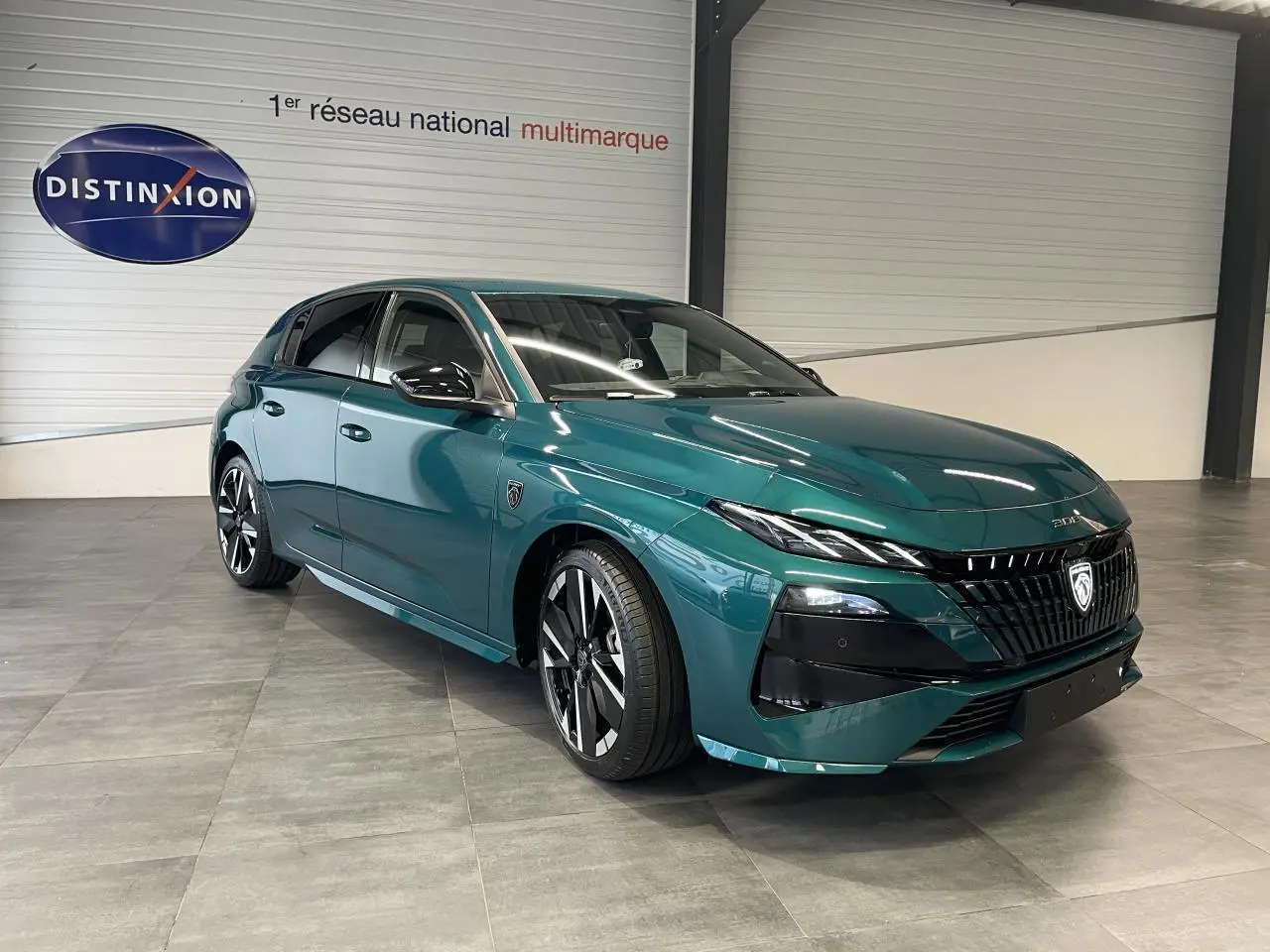 PEUGEOT 308 2026 bleu Lagoa en 3/4 avant droit avec calandre noire et jantes alliage bi-tons visibles en intérieur showroom.