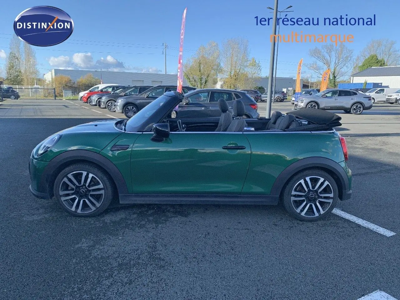 Profil côté gauche d'une MINI Cabriolet F57 2023 British Racing Green avec capote ouverte sur parking.