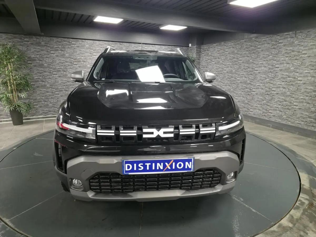 Vue avant d'un Dacia Duster 1.6 Hybrid noir nacré avec calandre moderne et plaque Distinxion en intérieur showroom.