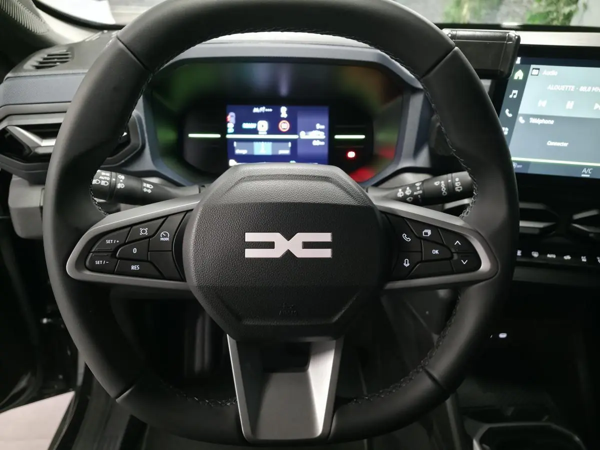 Vue rapprochée du volant noir du Dacia Duster 2025 avec commandes intégrées et tableau de bord digital en arrière-plan.