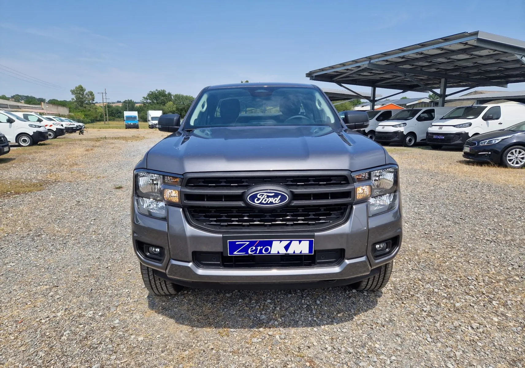 Vue frontale d'un Ford Ranger Super Cabine gris carbone, avec calandre noire et phares allumés, sur un parking extérieur.