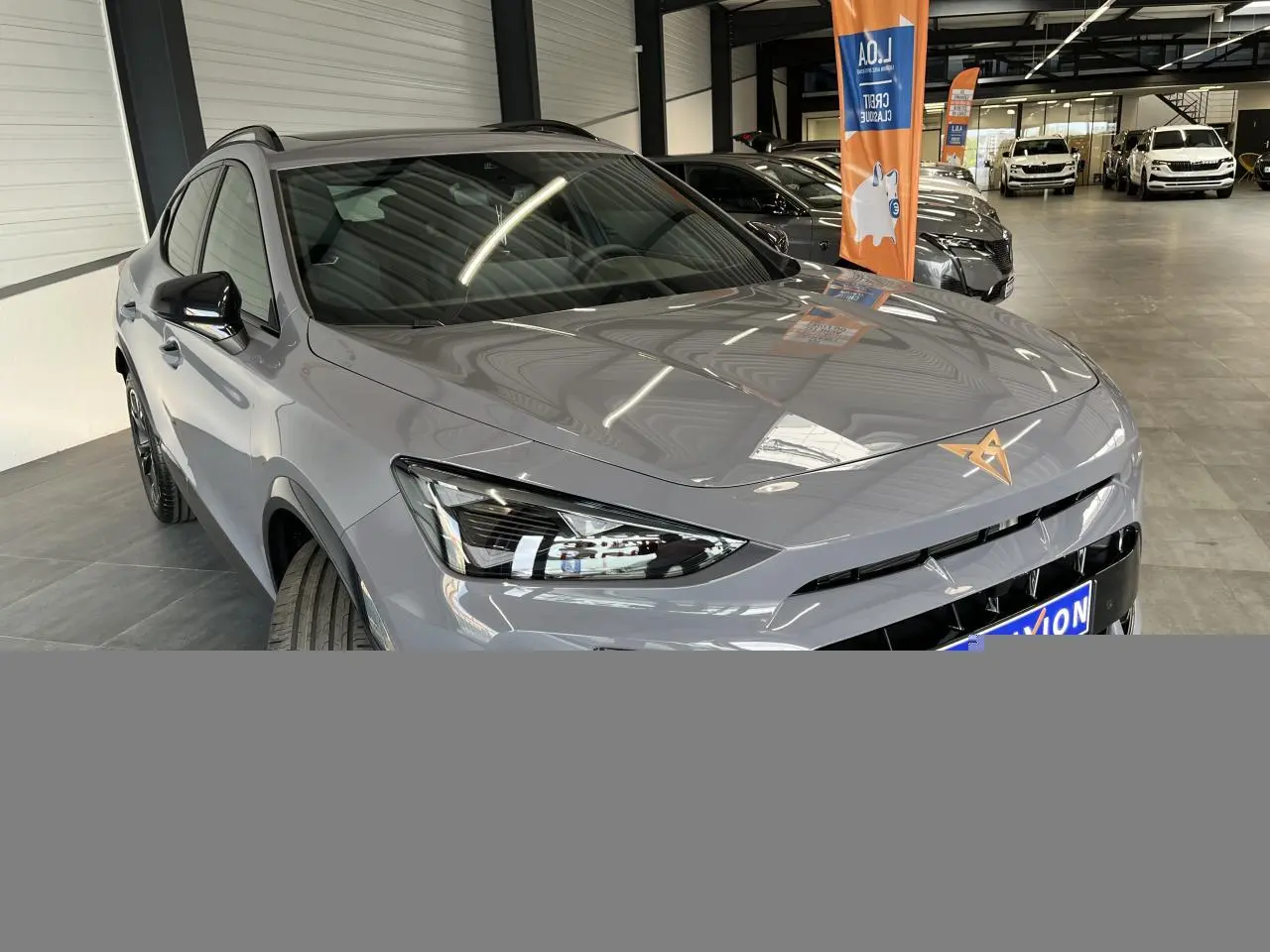 Vue 3/4 avant d'une CUPRA Formentor gris clair avec logo cuivré et rétroviseurs noirs dans un showroom.