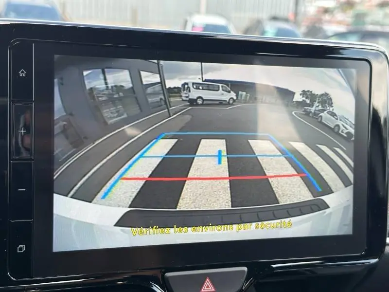Vue de la caméra de recul d'une Toyota Yaris 116h Dynamic blanche, montrant un passage piéton et un parking en arrière-plan.