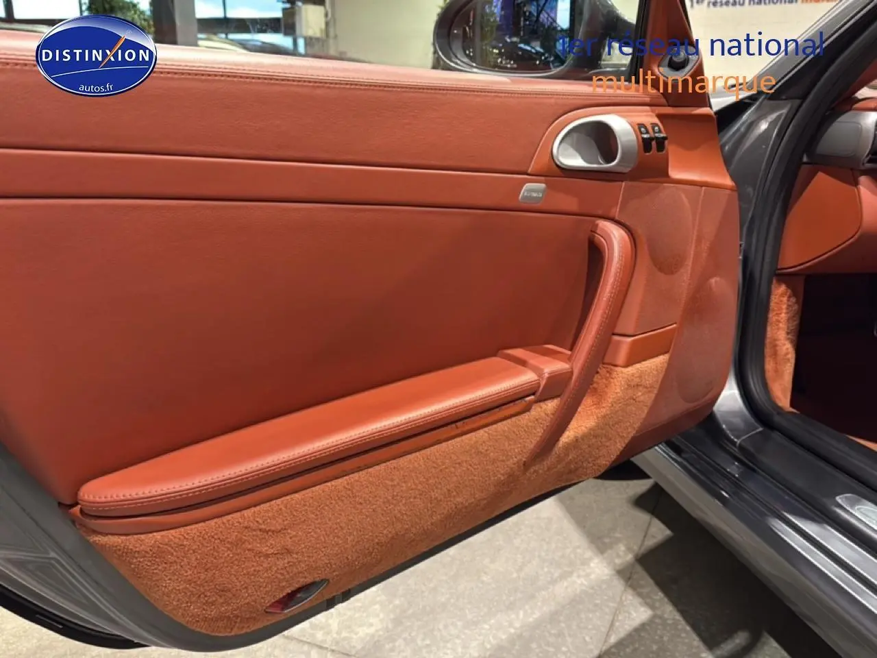 Vue intérieure côté gauche de la porte ouverte d’une Porsche 911 (997) Carrera PDK gris, avec garniture cuir marron et moquette assortie.