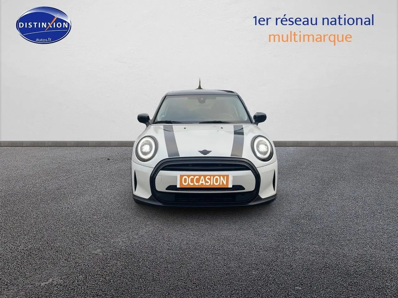 Vue frontale d'une MINI blanche nacrée 2023 avec bandes noires sur le capot et phares LED allumés.